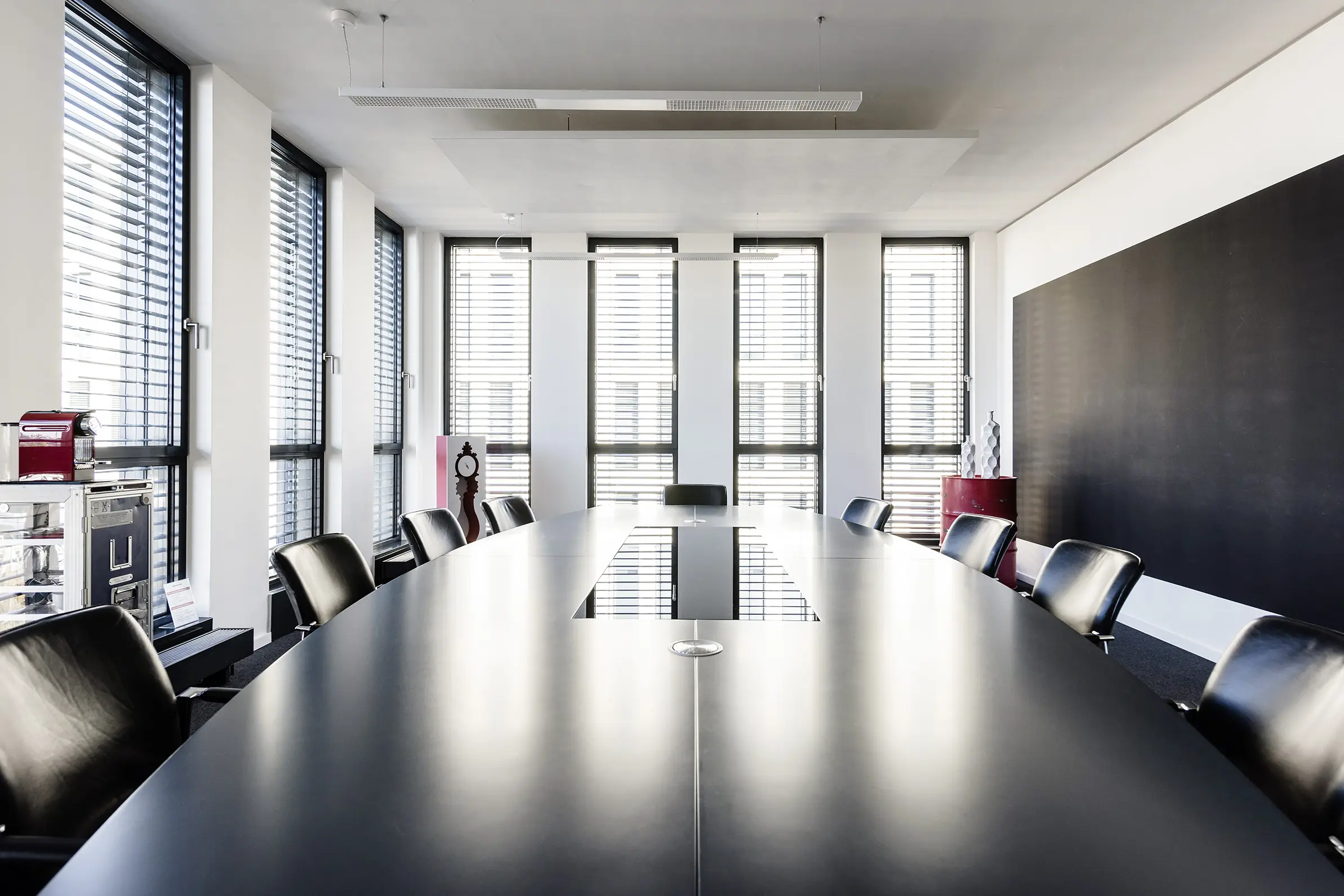 Langer maßgefertigter Konferenztisch in Schwarz mit Lederstühlen, großer dunkler Wandfläche und Fensterfronten mit Jalousien