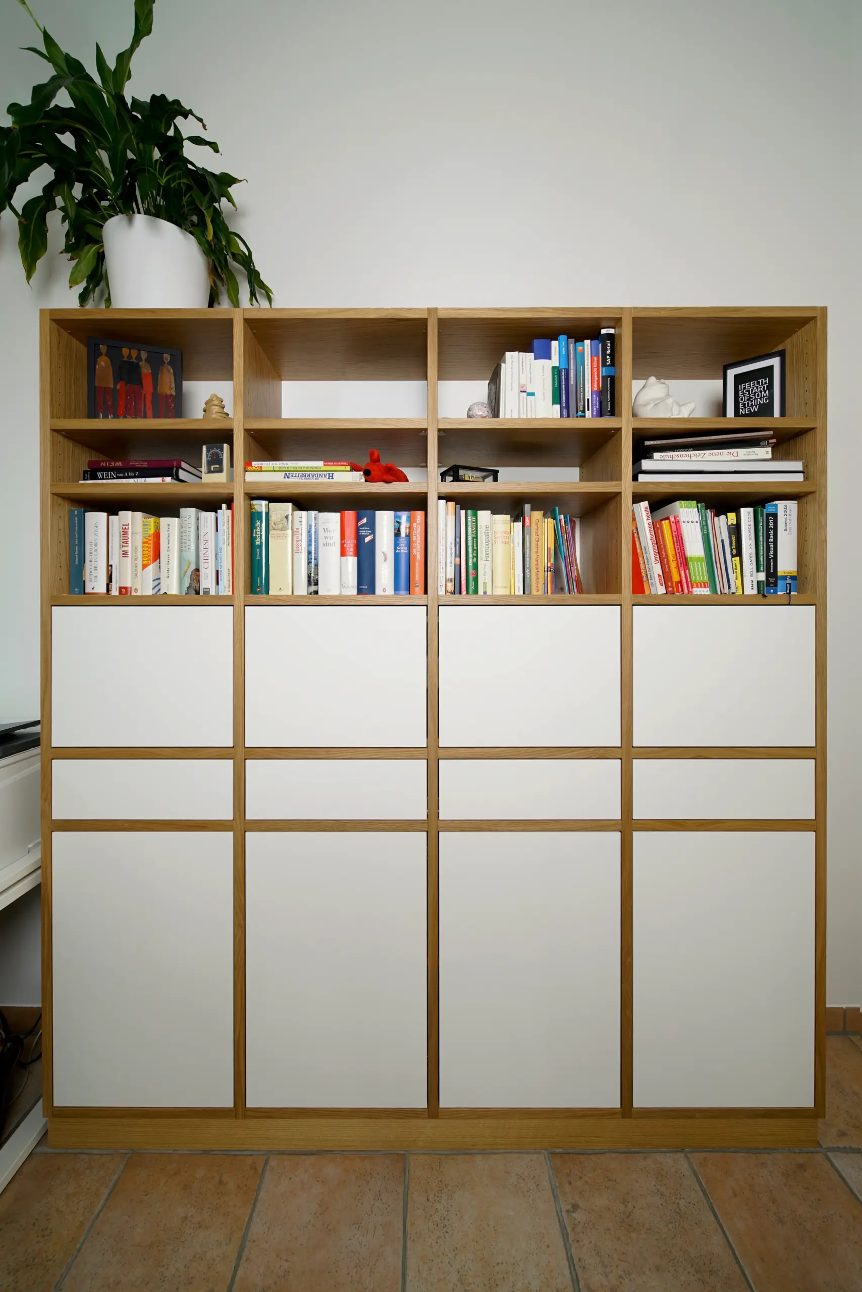 Großes Einbauregal aus Eiche mit weißen Fronten, offenen Fächern für Bücher und Dekoration sowie einer Zimmerpflanze auf dem oberen Regalabschluss