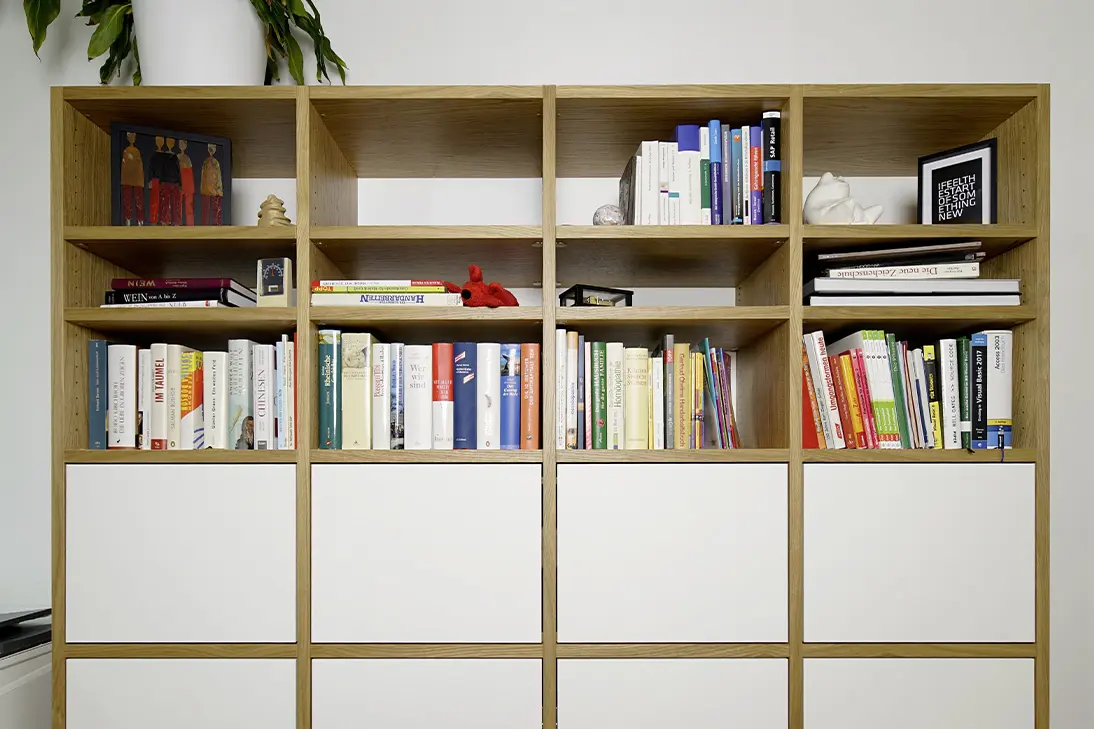 Großes Einbauregal aus Eiche mit weißen Fronten, offenen Fächern für Bücher und Dekoration sowie einer Zimmerpflanze auf dem oberen Regalabschluss