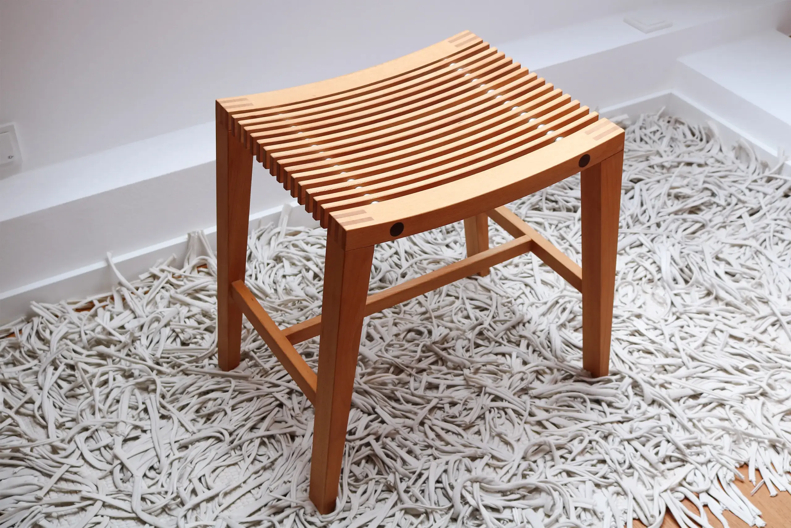 Handgefertigter Hocker aus Buchenholz mit Sitzfläche aus schmalen Holzleisten auf weißem Hochflorteppich