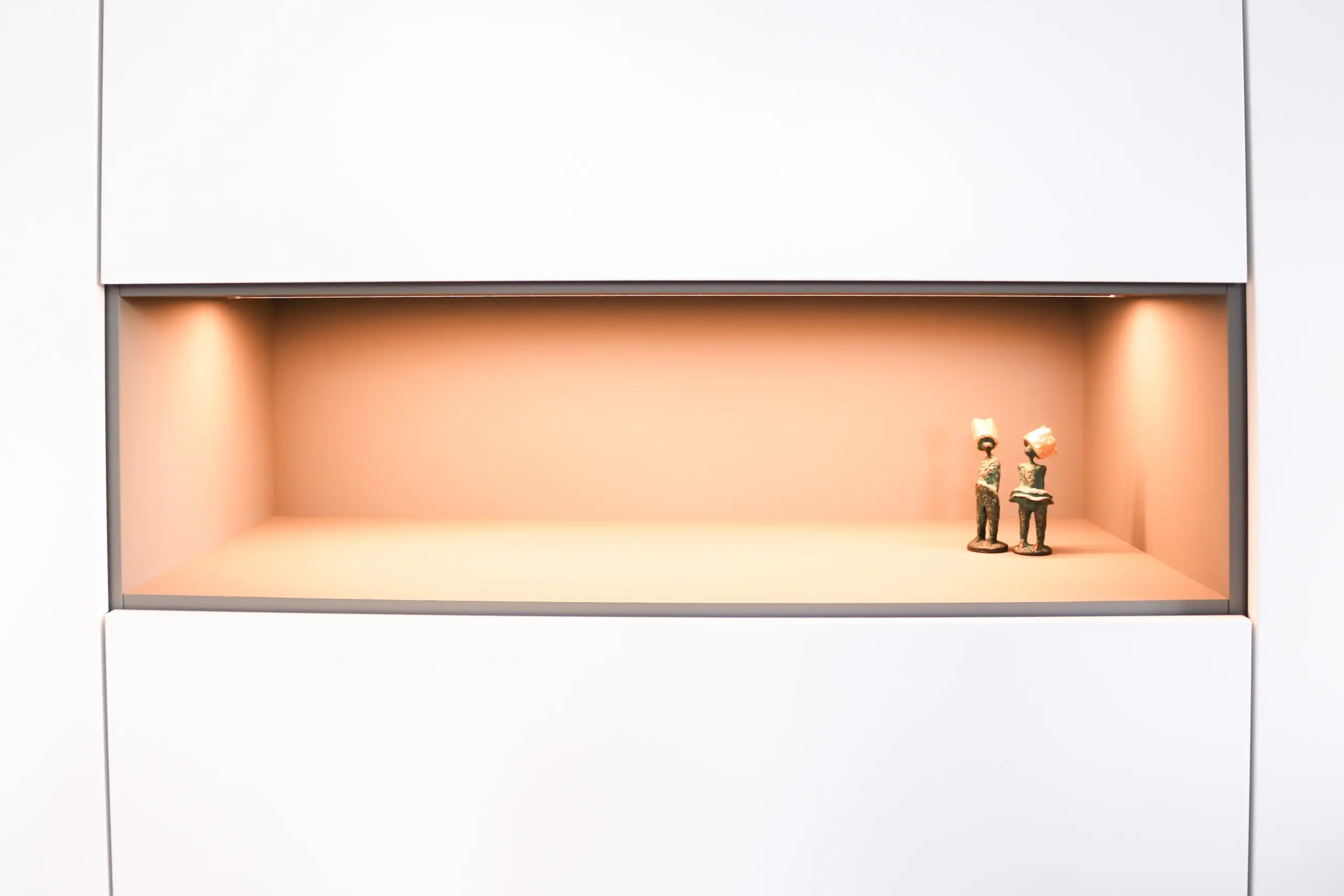 Detailansicht eines warm beleuchteten, in das weiße Sideboard integrierten Präsentationsfachs mit kleinen Figuren