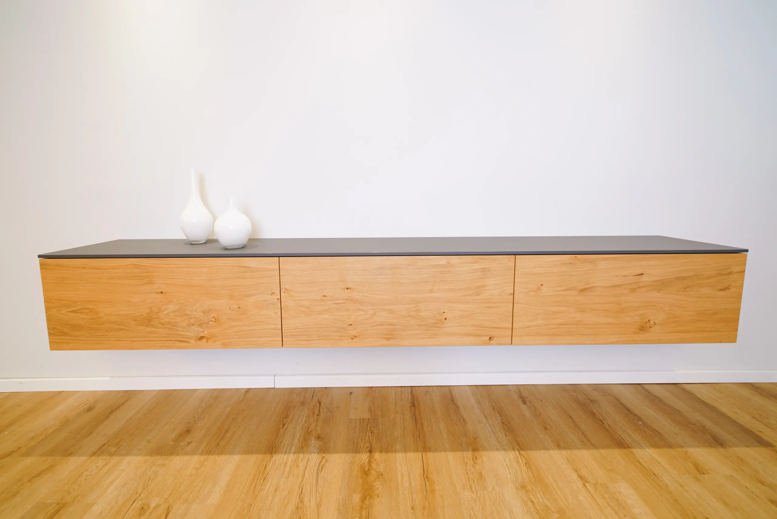 Schwebendes Sideboard aus Holz mit drei grifflosen Fronten und dunkler Abdeckplatte an einer weißen Wand