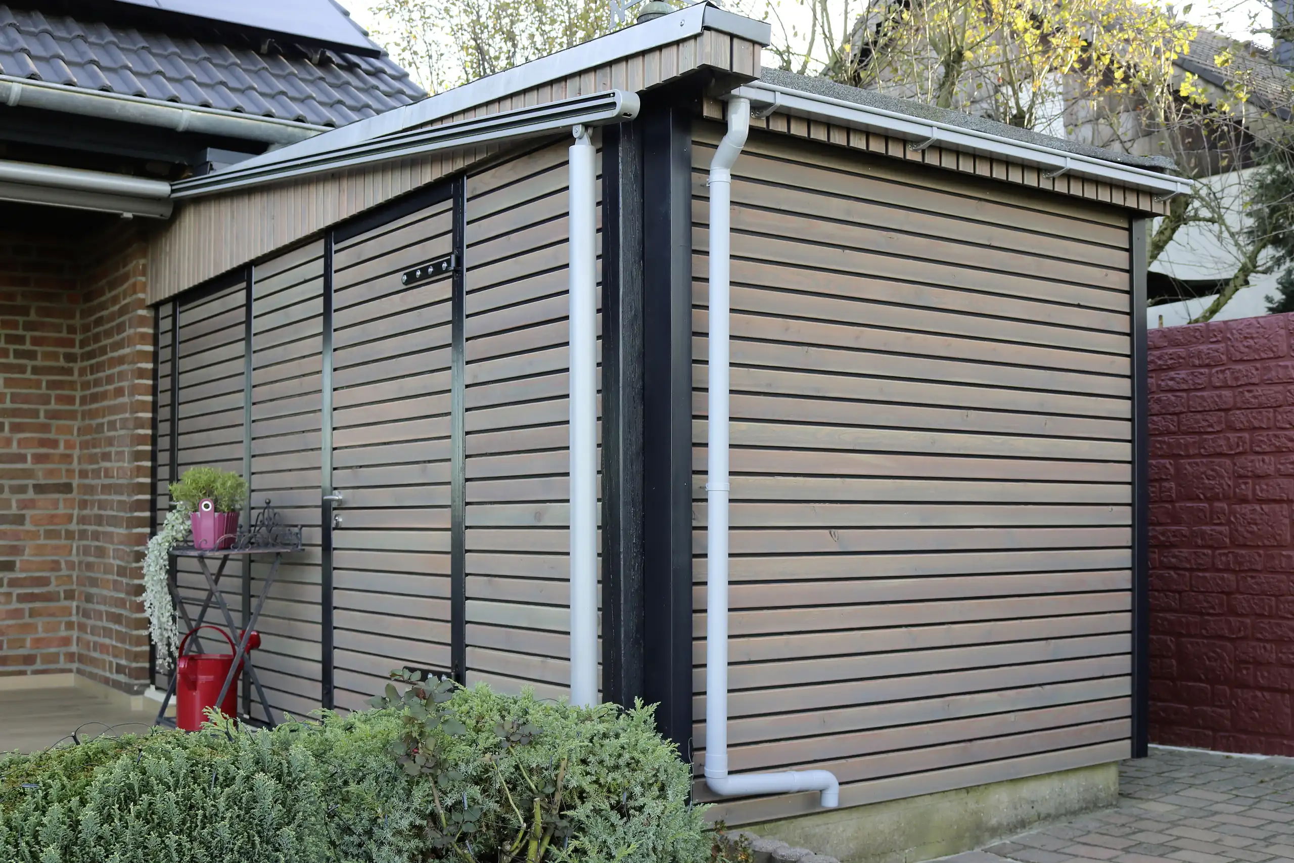 Gartenhütte mit grau lasierter Holzfassade, schwarzen Holzpfosten und grauem Regenfallrohr