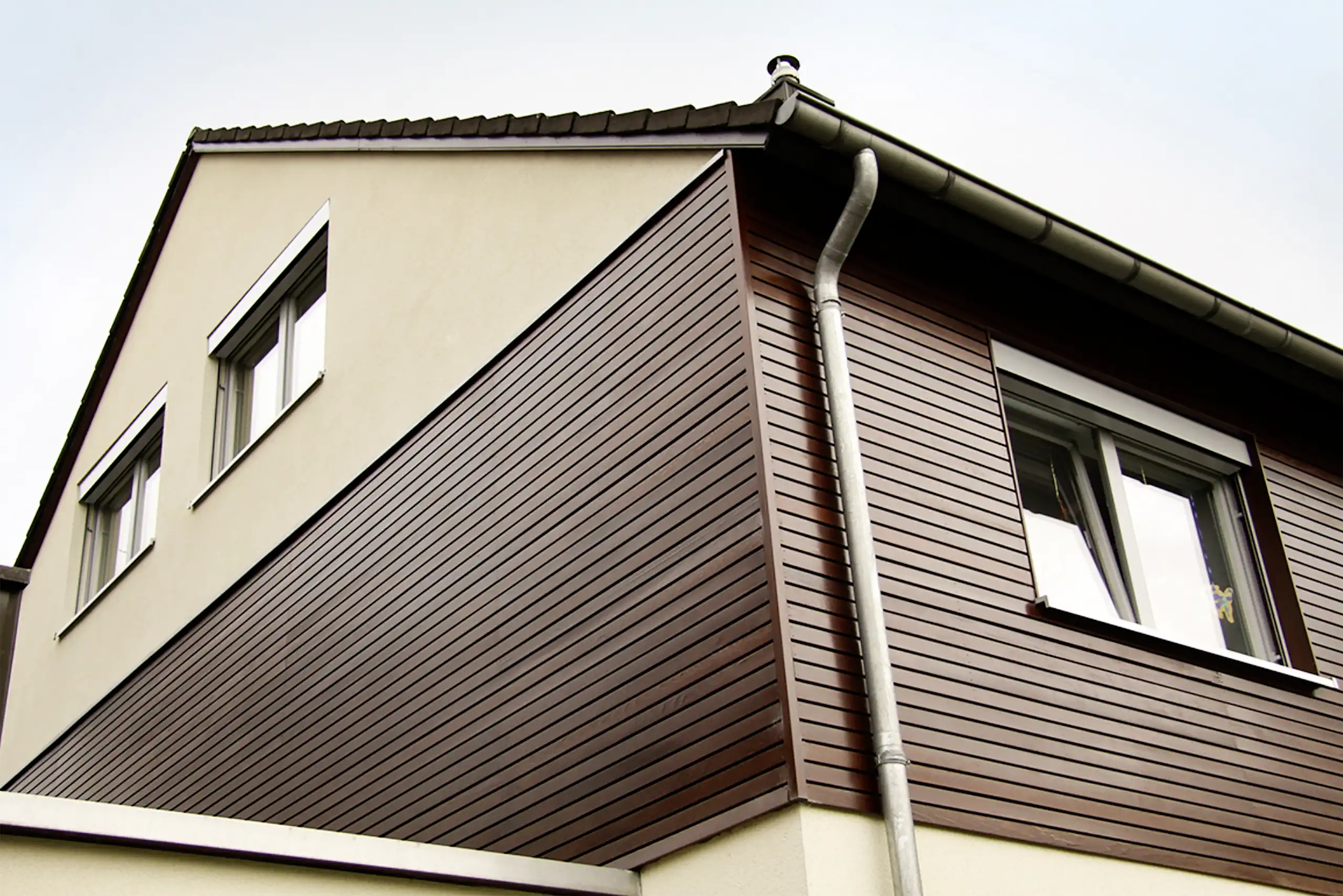 Außenansicht eines Wohnhauses mit dunkelbrauner Holzfassade, Fenstern und Regenrinne unter dem Dachüberstand