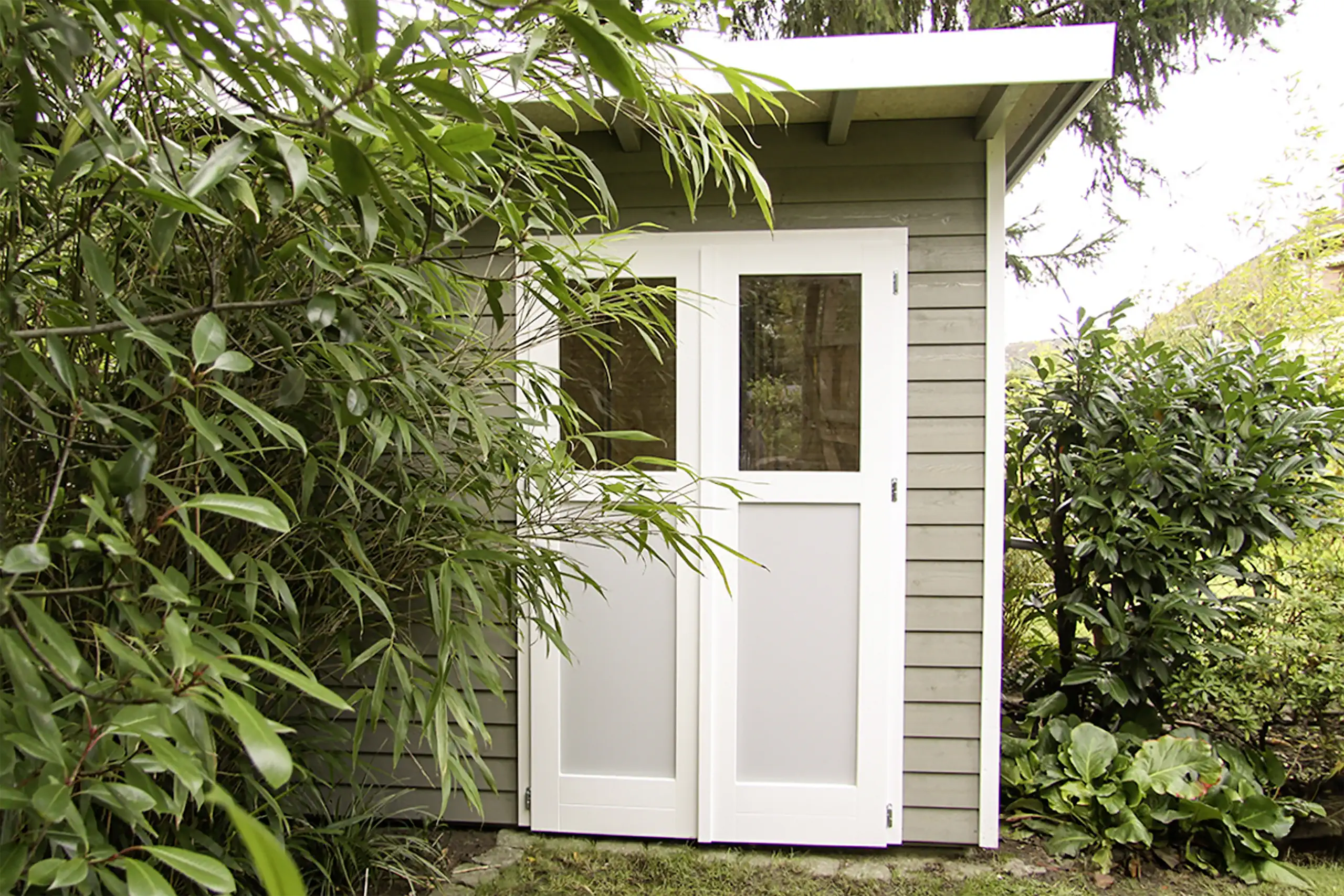 Moderne Gartenhütte mit grauer Holzfassade, weißen Doppeltüren mit Glaseinsätzen und dichter Bepflanzung