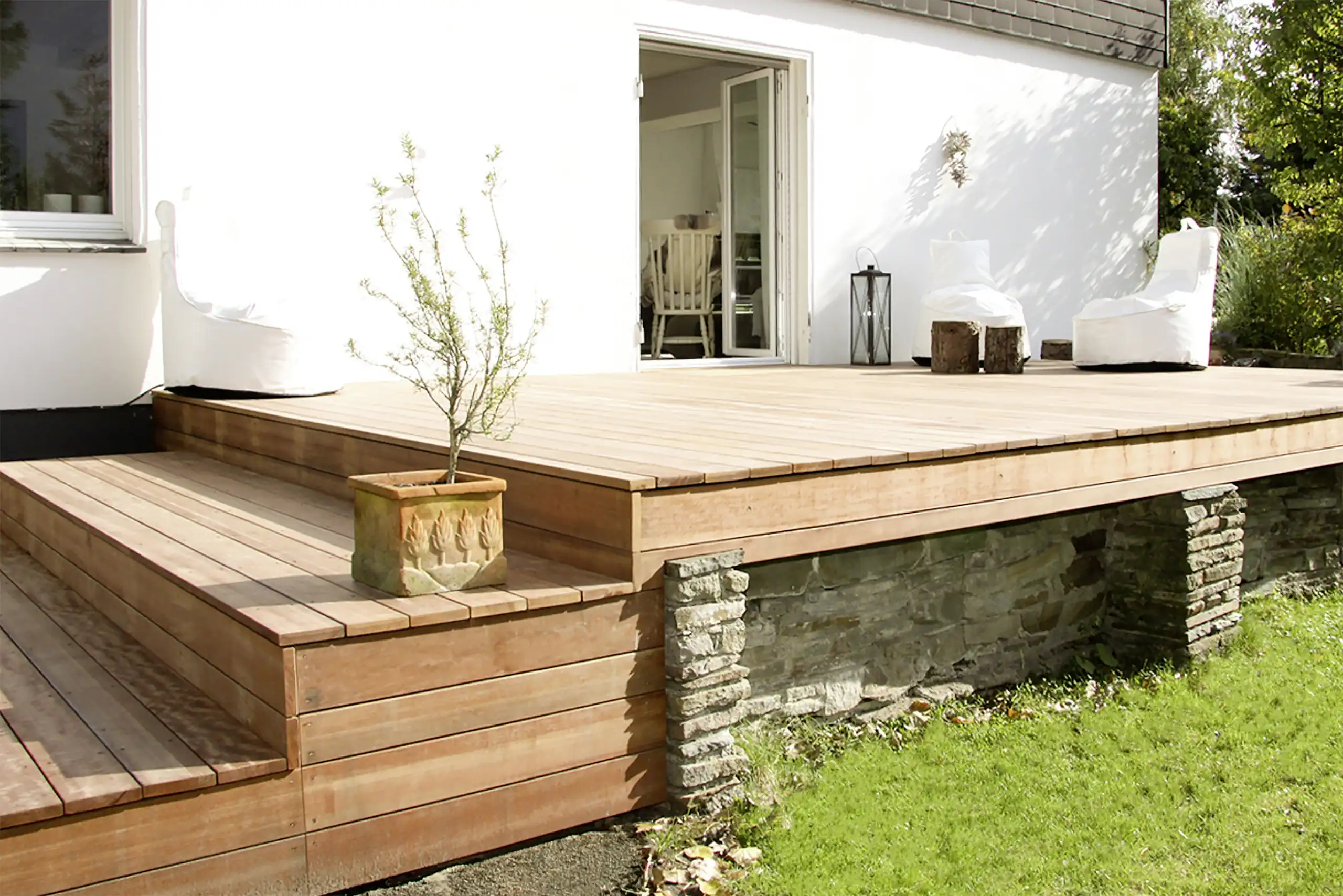 Großzügige, mehrstufige Holzterrasse mit weißen Lounge-Sesseln, Natursteinmauer und Blick in den Garten