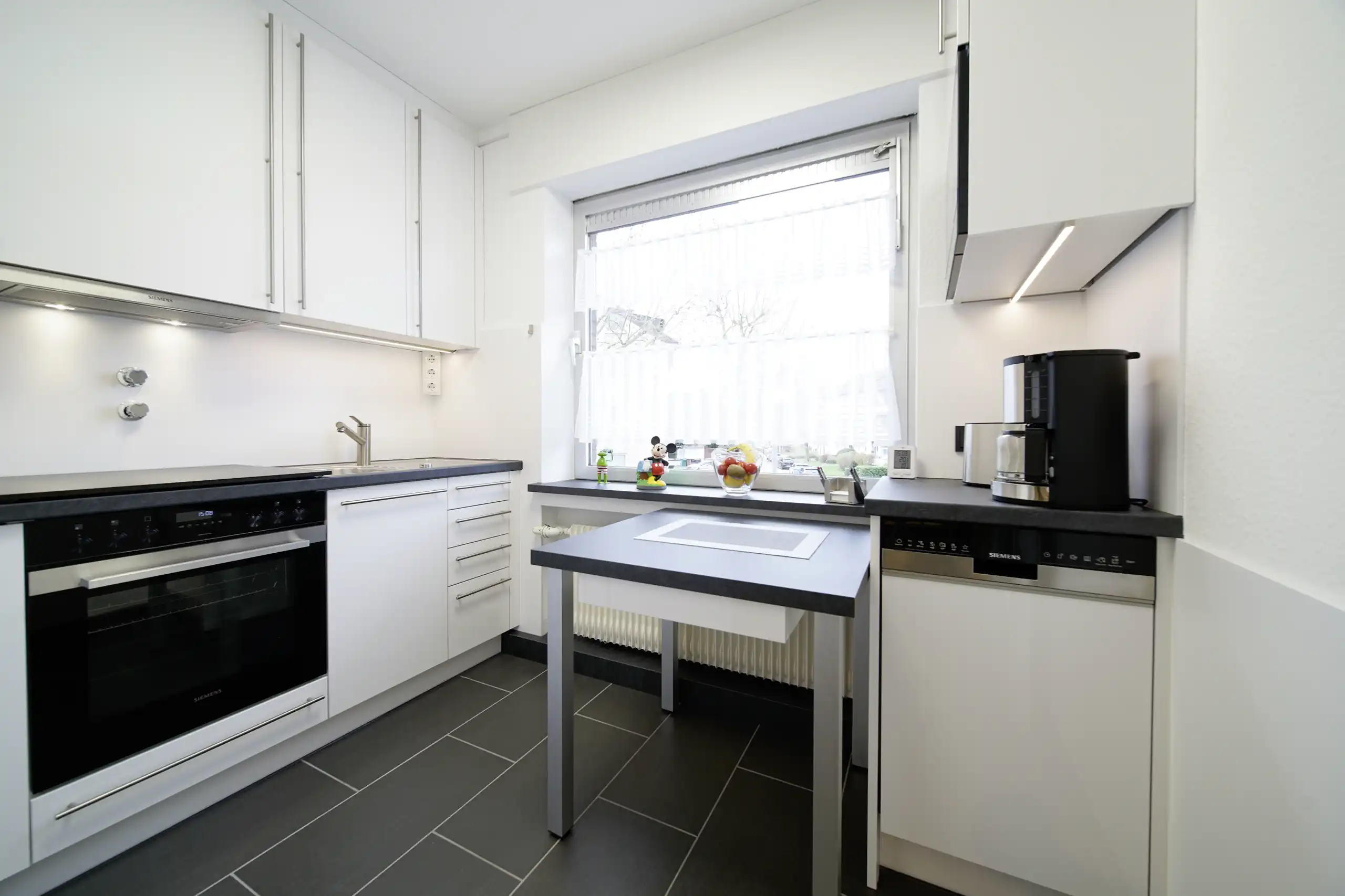 Kleine weiße Einbauküche mit dunkler Arbeitsplatte, Esstisch, Backofen, Geschirrspüler und Fenster