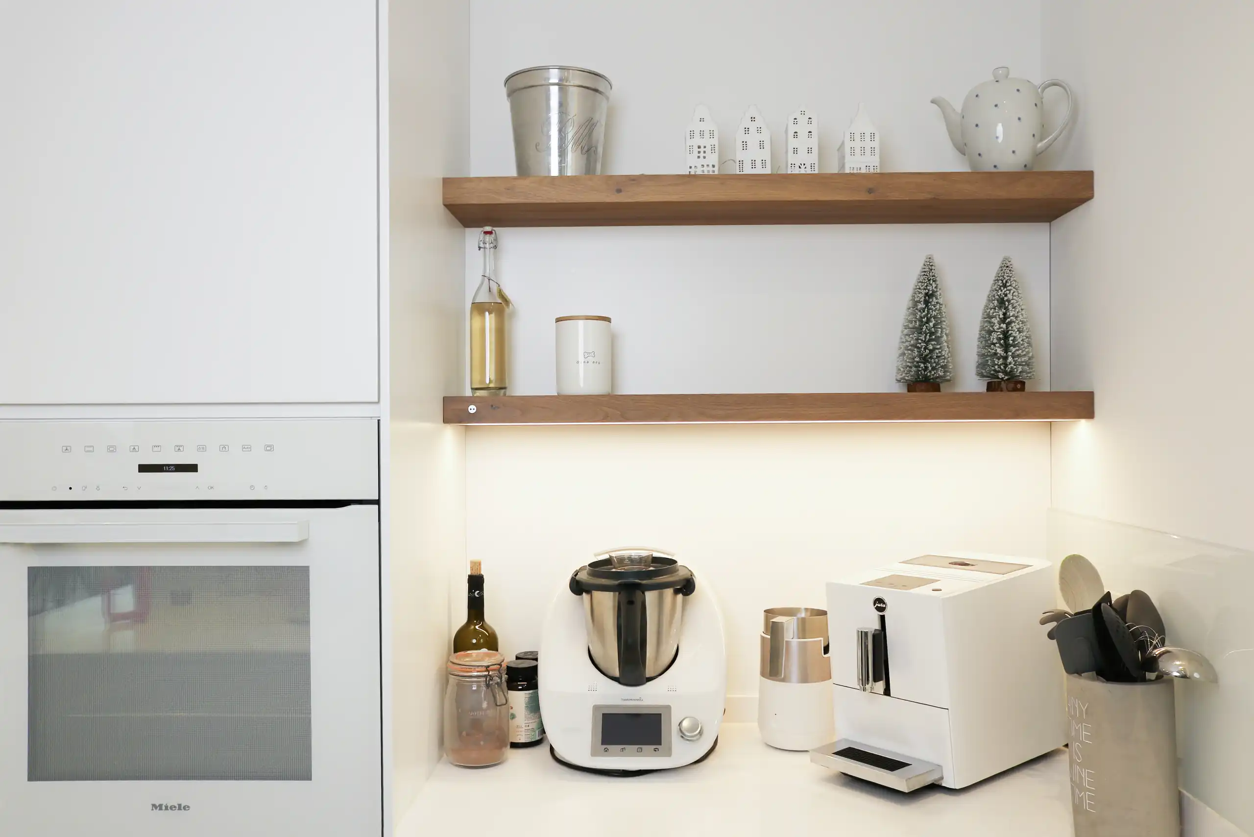 Arbeitsbereich einer weißen Küche mit Küchenmaschine, Kaffeevollautomat und beleuchteten Holzregalen