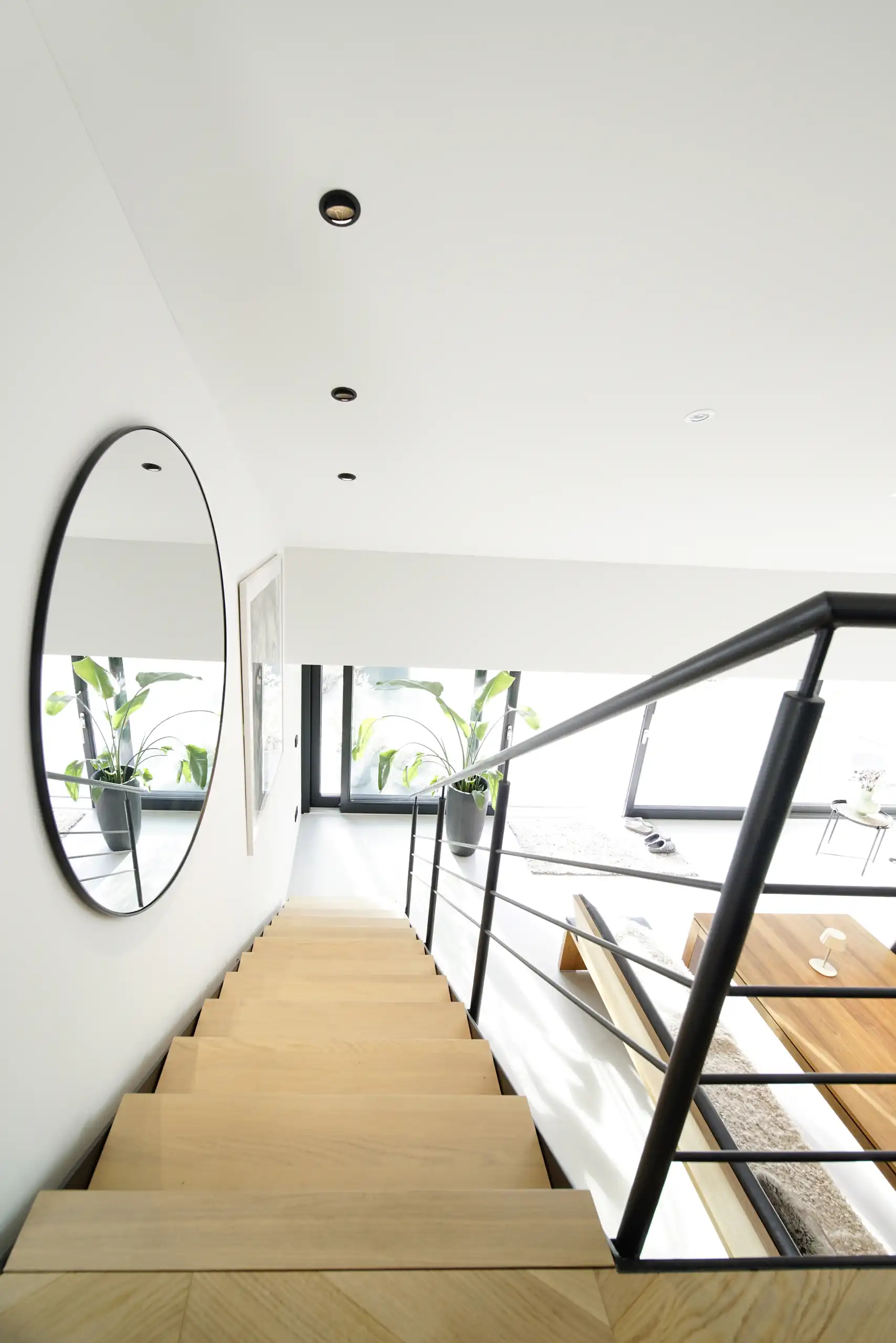 Blick über eine Treppe mit hellen Holzstufen und schwarzem Stahlgeländer in einen lichtdurchfluteten Wohnraum mit großem Rundspiegel