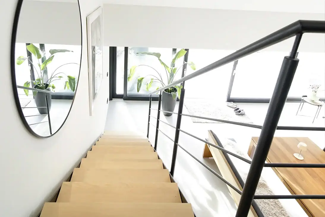 Blick über eine Treppe mit hellen Holzstufen und schwarzem Stahlgeländer in einen lichtdurchfluteten Wohnraum mit großem Rundspiegel
