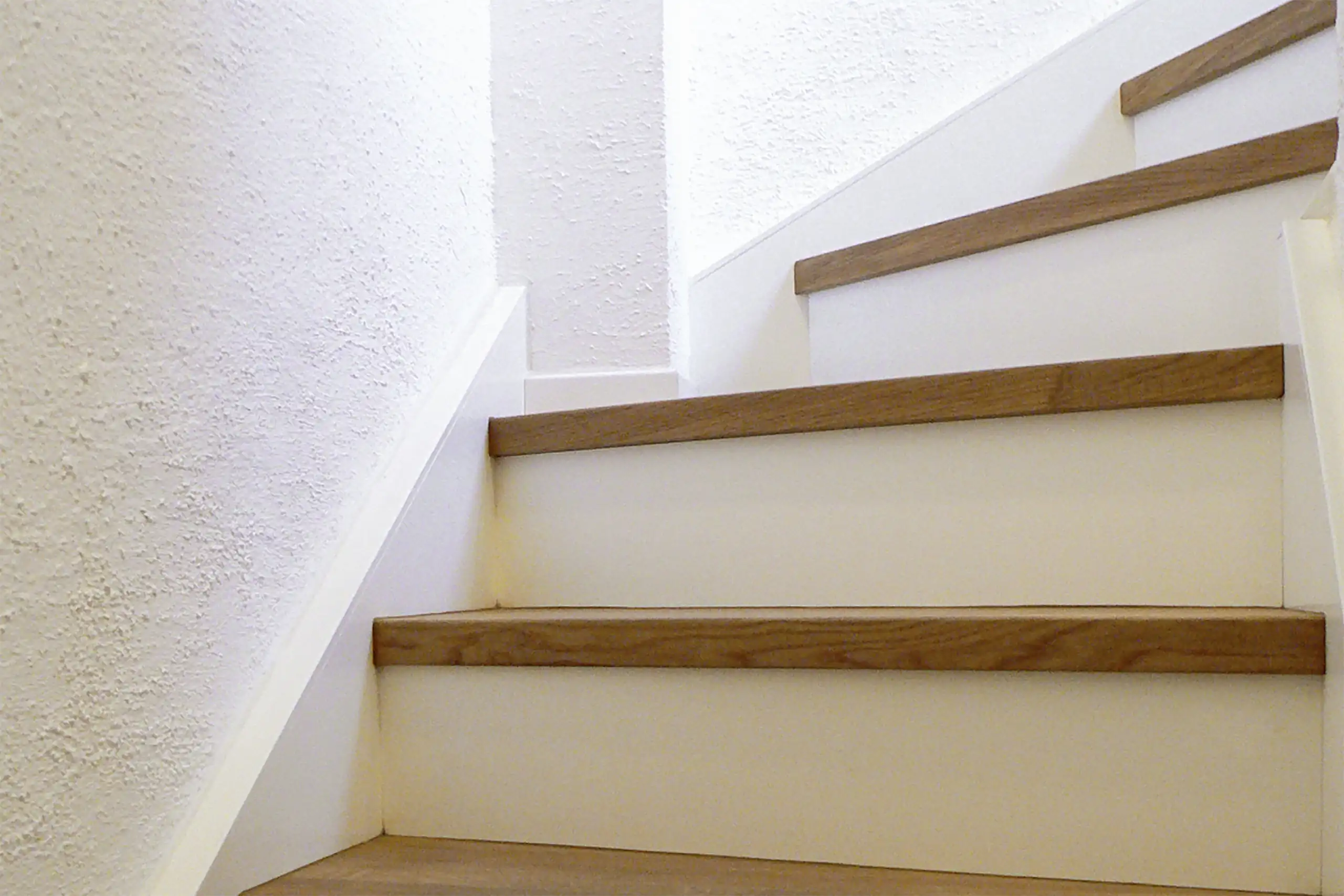 Aufwärts führende Treppe mit dunkel geölten Holzstufen, weißen Setzstufen und strukturierter weißer Wand