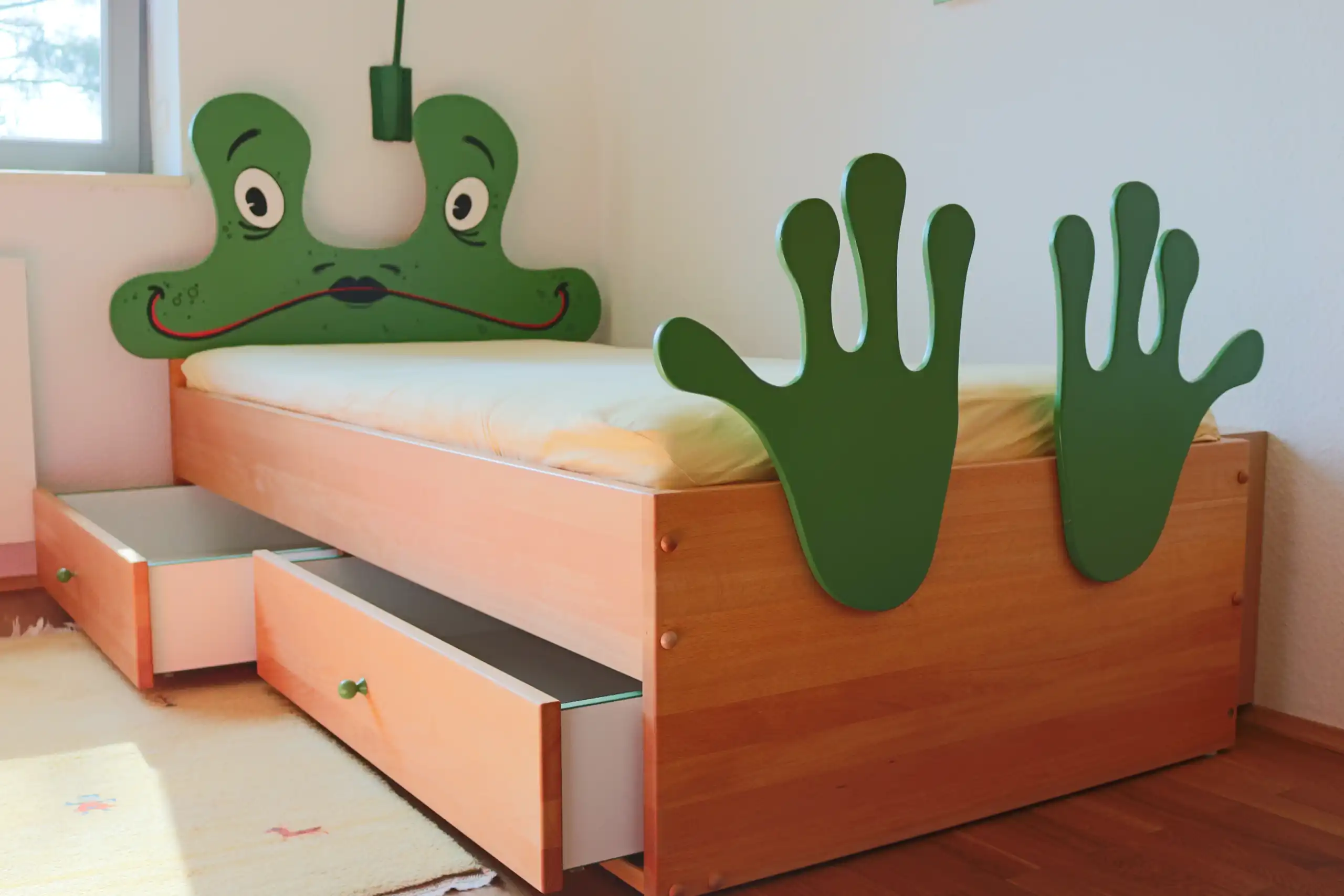 Kinderbett aus hellem Holz mit grünem Frosch-Kopfteil, froschförmigen Seitenteilen und ausgezogenen Bettkästen