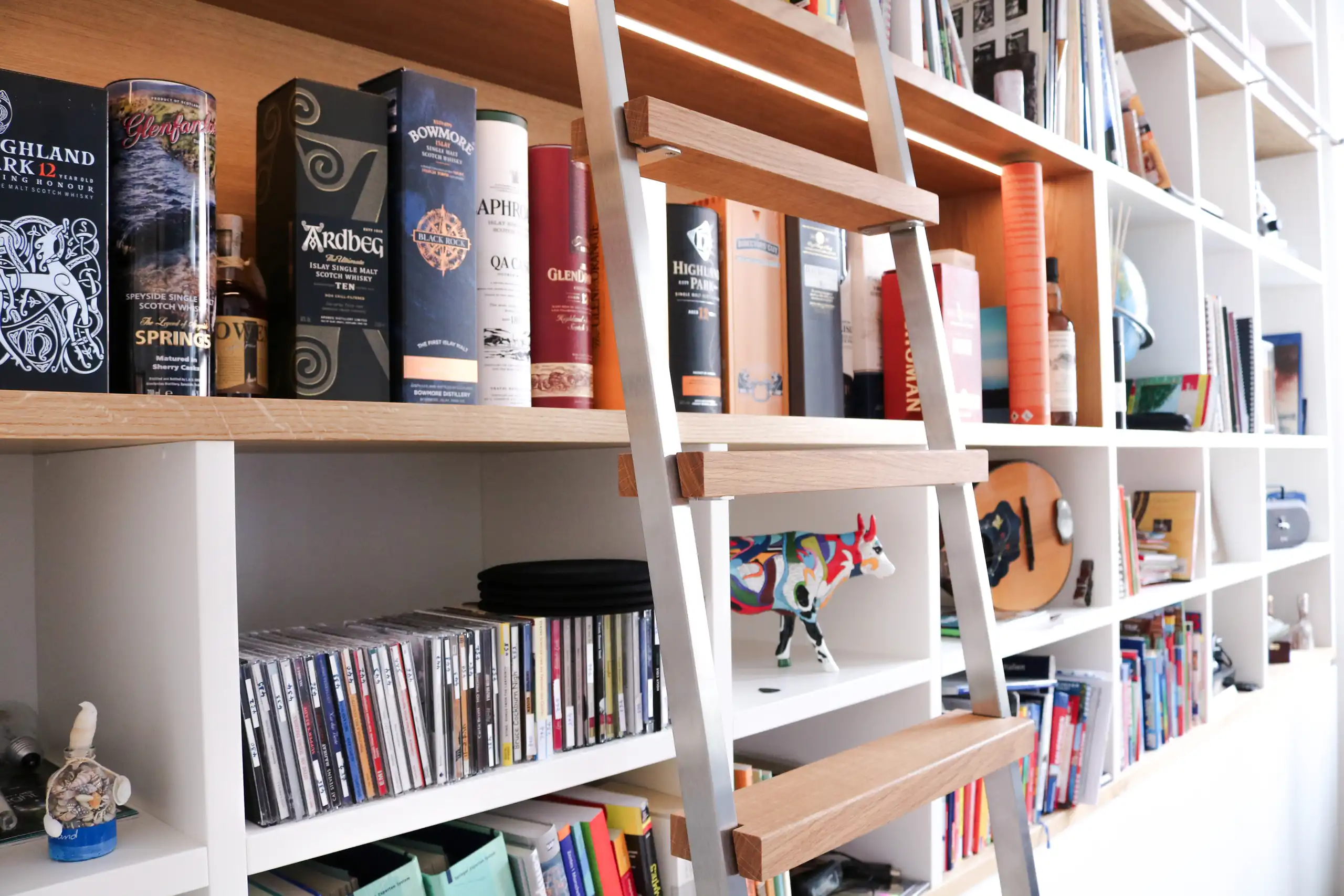 Maßgefertigte Bibliothek mit weißen Regalfächern, Holzablagen, integrierter Beleuchtung und Leiter auf Schienensystem