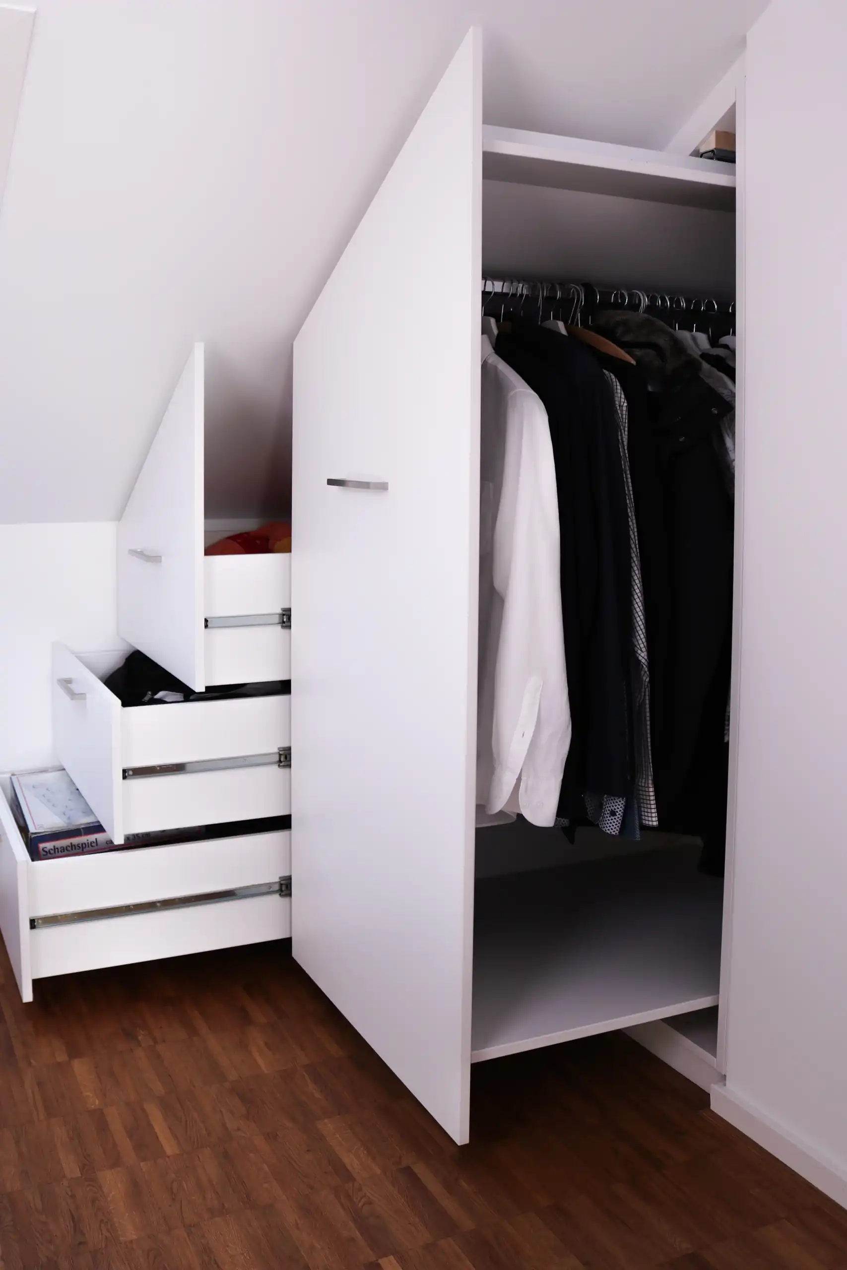 Geöffneter weißer Einbauschrank unter der Dachschräge mit Kleiderstange, Schubladen und passgenauem Stauraum