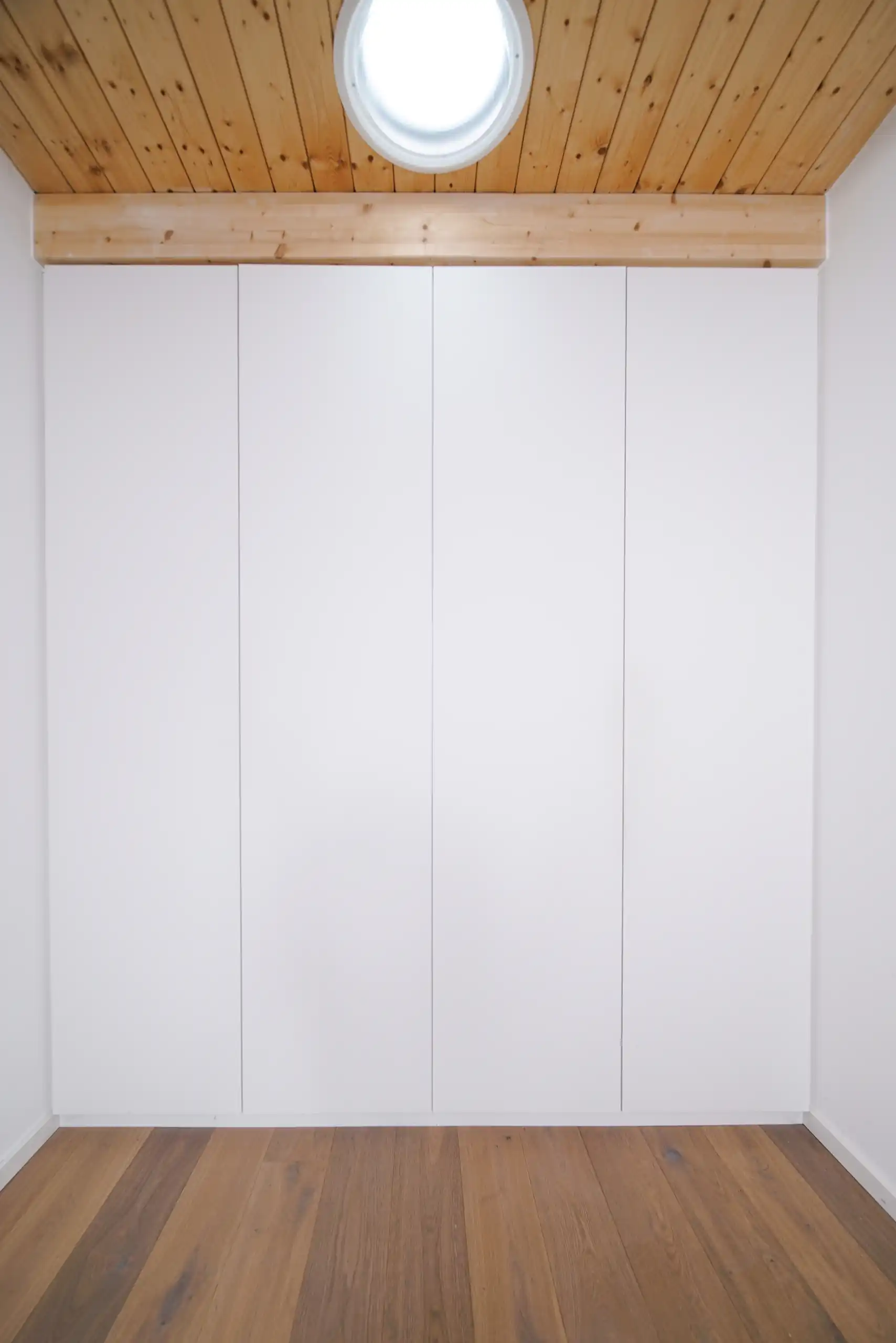 Weißer Einbauschrank mit grifflosen, bündigen Fronten unter einer Holzdecke mit rundem Dachfenster