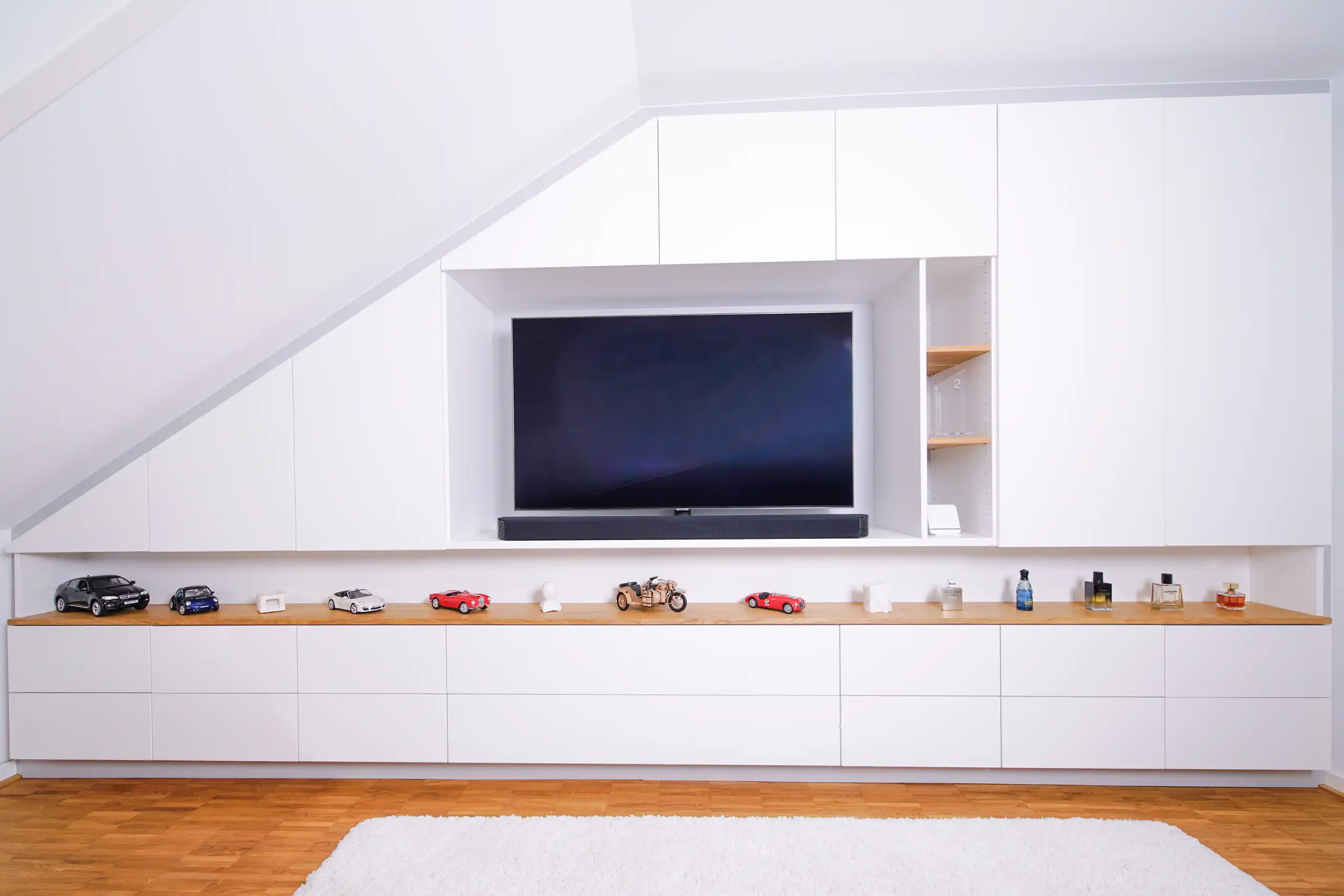 Passgenaue weiße Einbaulösung unter der Dachschräge mit Fernseher, Soundbar, durchgehender Holzablage und großzügigem Stauraum