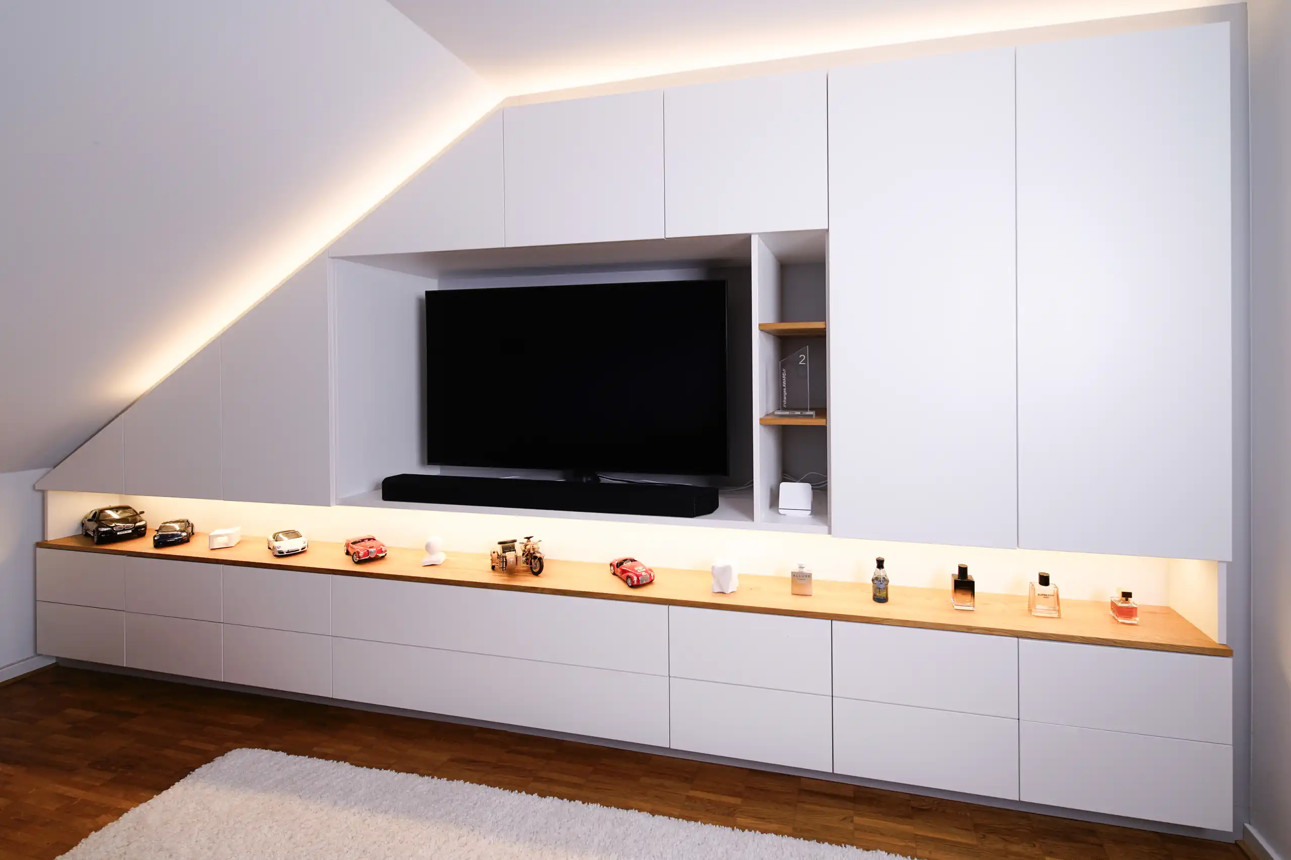 Maßgefertigte weiße Einbauschrankwand unter einer Dachschräge mit integriertem TV-Fach, Holzablage und indirekter Beleuchtung