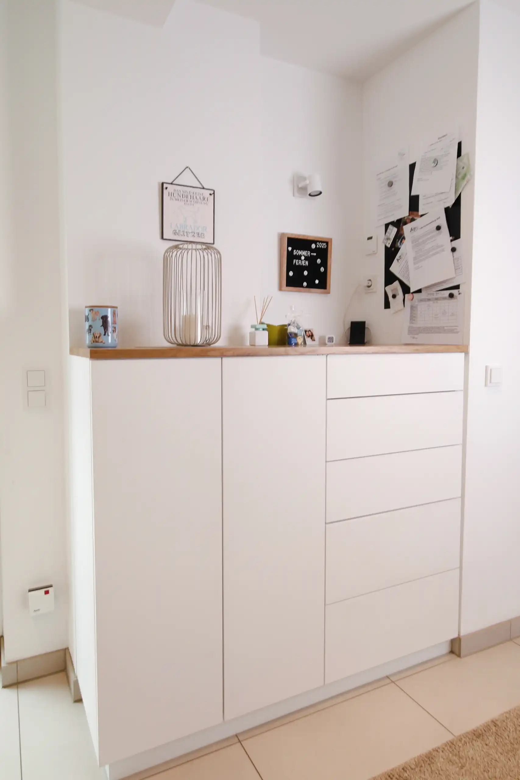 Weiße Kommode im Eingangsbereich mit Holzauflage, zwei Schranktüren, mehreren Schubladen sowie einer Pinnwand und Dekoration an der Wand.